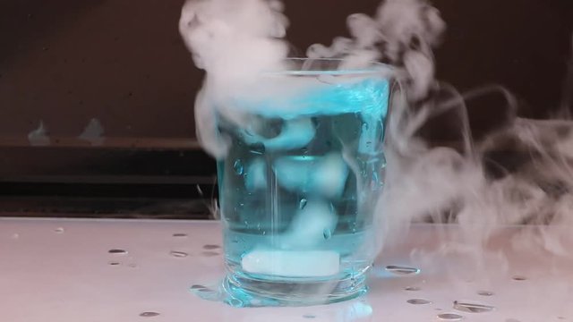 Dry ice in a glass of blue water and engender white smoke on white floor with black background.