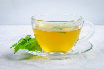 Leaves of fresh green nettle and a clear glass cup of herbal nettle tea on a gray concrete table.