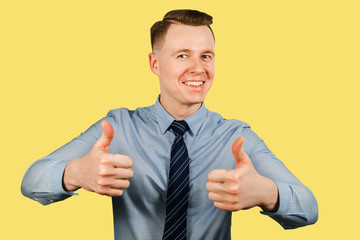 Young businessman dressed in blue shirt and tie shows thumbs up and smiles, isolated on yellow background
