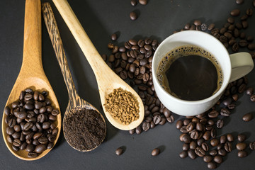 Hot coffee in white ceramic glass and coffee beans, coffee powder and instant coffee are placed in a wooden spoon placed on a black table top.