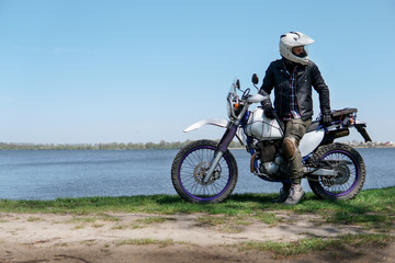 Young stylish man sit on classic retro off road track motorcycle on the beach, outdoor portrait, posing, in lather jacket and Sunglasses, travel active lifestyle concept, ocean, sea, lake, river
