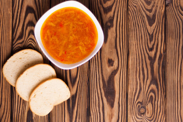 One full white ceramic bowl of fresh hot vegetable borscht and three pieces of bread on old weathered rustic wooden table. Top view
