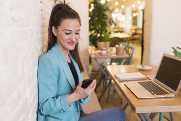 portrait of a young beautiful woman using mobile phone in a restaurant. She is smiling. Modern life of a blogger with computer laptop, tablet, notebook and coffee on table. Casual clothes. Lifestyle