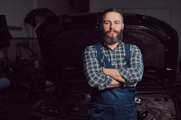 Obraz na płótnie Canvas A bearded expert mechanic dressed in a uniform, standing with crossed arms against a car in the garage.