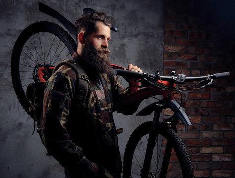 A handsome bearded traveler, dressed in a trendy camouflaged jacket, standing with sports bicycle on a shoulder in a studio, isolated on a dark background.