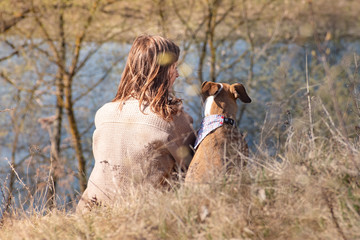 Girl and dog in bandana sit in grass near river on the hill. Young female with staffordshire...