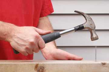 Closeup view of man hammering nail with house siding in background.