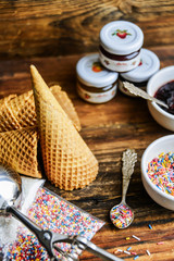 A Kit For Making Ice Cream: Spoon For Ice Cream, Ice Cream Cones, Jam, Topping, Chocolate, Sprinkles, Coconut Shavings. On Wooden Background. The Concept Of A Set, Surprise, Gift Idea