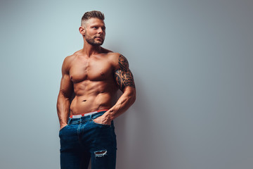 A handsome shirtless tattooed bodybuilder with stylish haircut and beard, wearing sports shorts, posing in a studio. Isolated on a gray background