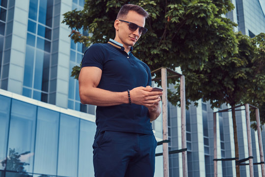 Young handsome fashionable man with a stylish haircut in sunglasses, dressed in a black t-shirt and pants, standing in the modern city against a skyscraper.