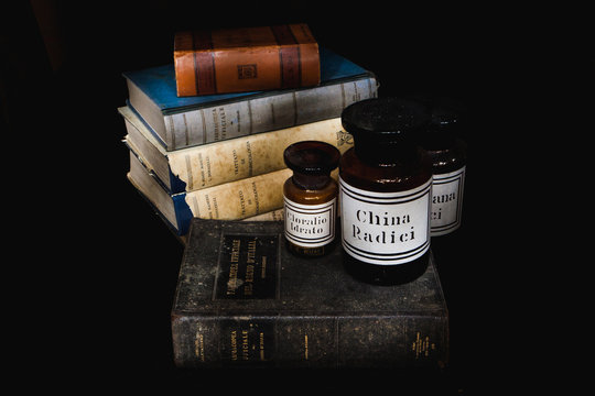 Old books (Italian Pharmacopeia and Pharmacology books) and old glass pharmacy vases (Valerian, Chloral, Quinine). Light painting picture, black background.