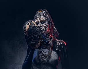 Make-up concept. Portrait of a scary African shaman female with a petrified cracked skin and dreadlocks, holds a traditional mask on a dark background. Make-up concept.