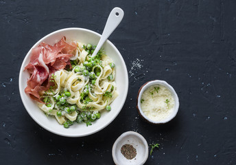 Fettuccine pasta with creamy sauce, green peas and prosciutto on dark background, top view