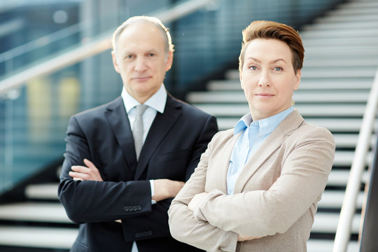 Elegant mature businessman and businesswoman looking at camera with crossed arms by staircase of airport lounge