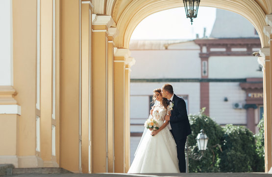 Beautiful wedding couple embrace standing near the pillars, and groom kissing on the cheek his bride. Outdoors
