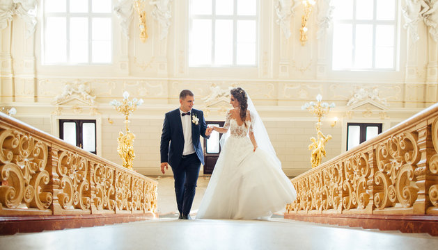 Beautiful wedding couple climbing up a gorgeous stairs holding hands and looks to each other. Indoors..