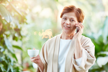 Contemporary female speaking on smartphone while drinking coffee inside orangery