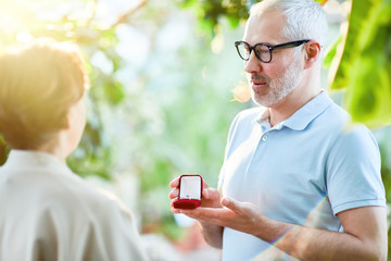 Aged man with grey hair showing diamong ring in red velvet box to his wife or sweetheart in orangery