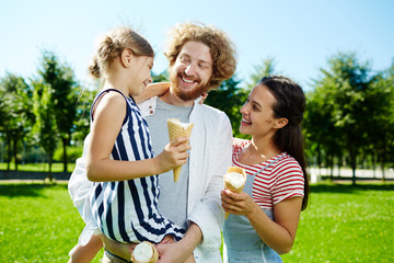 Happy little girl and her parents with ice creams talking and enjoying sunny day in park