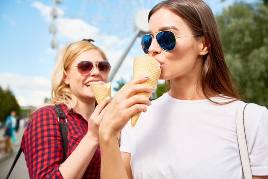 Two stylish girls in sunglasses eating tasty ice cream in amusement park on background of blue sky and ferris wheel