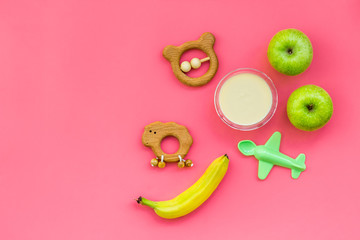 Fruit puree for baby. Jar with food, apple, banana, toys on pink background top view copy space