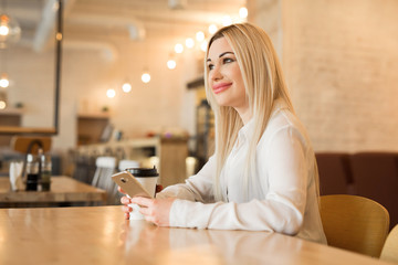 beautiful young girl in a white shirt with a cup of coffee and a phone in hands at a restaurant