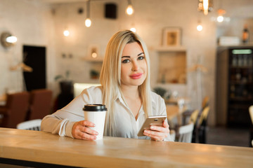 beautiful young girl in a white shirt with a cup of coffee and a phone in hands at a restaurant