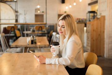 beautiful young girl in a white shirt with a cup of coffee and a phone in hands at a restaurant