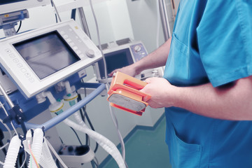 Male doctor with defibrillator electrodes in his hand sets the discharge parameters in front of the life support monitor - obrazy, fototapety, plakaty