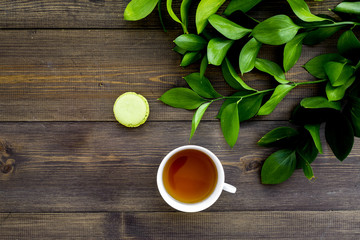 Summer mood, summer tea party. Cup of tea and sweets macarons near brignt greenery on dark wooden background top view copy space