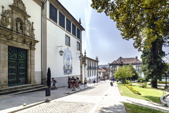 Guimaraes, Portugal. August 14, 2017: Facade Of The Catholic Church Called Nossa Senhora Do Carmo With A Mosaic Of Tiles With The Image Of The Virgin Mary And Carmen Garden With Young People Walking