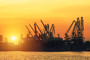 Fototapeta na wymiar A city landscape. Bright sunset in the seaport. A sunny path on the calm surface of the sea. Large silhouettes of loading cranes. Bright abstract background ideal for any design 