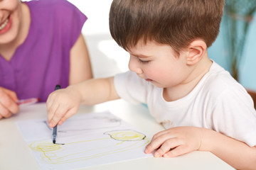 Close up shot of dark haired handsome male kid being focused at paper with picture, draws with great desire, unrecognizable female sits next to him, helps to make real masterpiece. Childhood