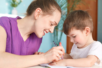 Horizontal shot of happy young smiling mother being glad to spend free time with her small son, draw something with great interest, enjoy togetherness, have good relationships. Leisure concept