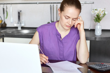 Cropped shot of pleasant looking female with no make up, dressed casually, being concentrated in documents, calculates family budget, works on portable laptop computer, sits in cozy kitchen.