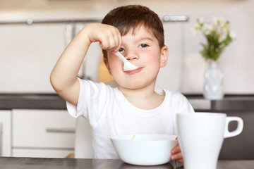Horiontal portrait of attractive male child eats delicious porridge with milk, dressed in casual white t shirt, has good appetite after walk outdoor with mum, sits at kitchen table with bowl and mug