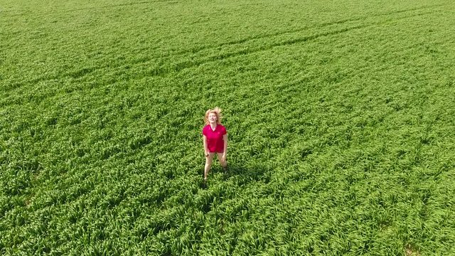 A beautiful young blonde girl is walking along the field of young wheat. A woman in a red T-shirt poses in front of the drony camera. He runs away