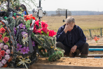 A gray-haired tired man or widower is sitting near a fresh grave in the cemetery and mourning over...