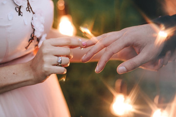 Wedding ceremony. Bride in a pink dress is wearing an engagement ring to the groom on the background of a bokeh of light bulbs. On her hand is a gold ring with a diamond close-up.