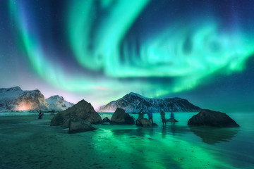 Green aurora borealis and photographers. Aurora. Northern lights in Lofoten islands, Norway. Starry sky with polar lights. Night landscape with aurora, sea, people, stones, sandy beach and mountains. - obrazy, fototapety, plakaty