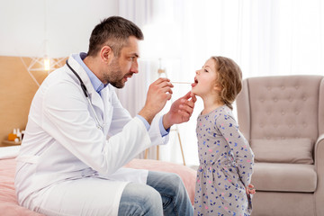 Open widely. Concentrated professional doctor examining his patient and looking in her throat