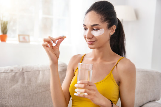 Helpful pills. Upbeat young woman sitting on the sofa and being about to take a vitamin pill while wearing under-eye patches