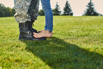 Fototapeta na wymiar Close up feet of soldier and daughter. Leather military boots and naked feet on the meadow.