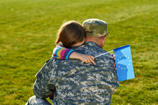 European soldier is hugging his little daughter. Back view, man in camouflage is embracing her little daughter with blue EU flag.