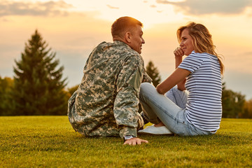 Romantic meeting of soldier and his girlfriend outdoor. Sitting on the lawn grass and talking,...