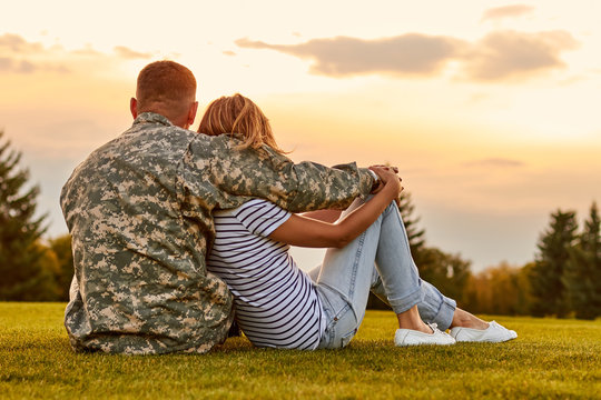 Happy couple looking at sunset. Back view, soldier and his wife sitting on the grass.