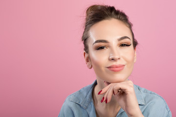 Portrait of cute elegant girl is looking at camera with joy while touching her chin. Isolated on pink background. Copy space in the left side
