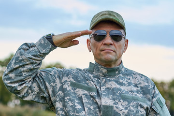 Sergeant in sunglasses saluting portrait. Severe strong soldier in spectacles saluting to officer.