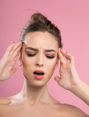 Fototapeta premium Awful headache. Portrait of upset girl is standing with closed eyes and touching her head while suffering from pain. Isolated on pink background