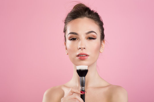 Perfect makeup. Portrait of naked young elegant woman is standing with professional brush. She is looking at camera confidently. Isolated on pink background